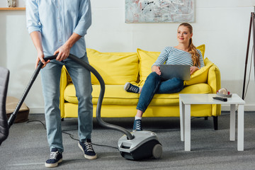 Fototapeta na wymiar Smiling girl with laptop looking at boyfriend with vacuum cleaner in living room
