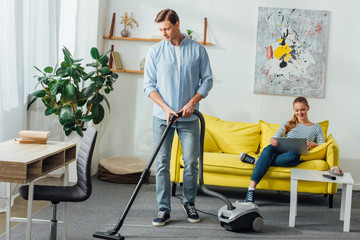 Man cleaning carpet with vacuum cleaner near smiling girl using laptop on couch at home