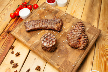 Juicy grilled marbled beef steaks lie on a wooden cooking board. Nearby a bunch of cherry tomatoes and sauces.