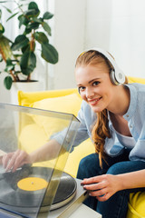 Smiling girl in headphones looking at camera near record player in living room
