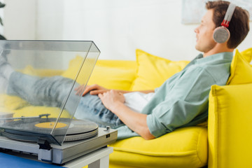Selective focus of record player on coffee table and man in headphones lying on couch at home