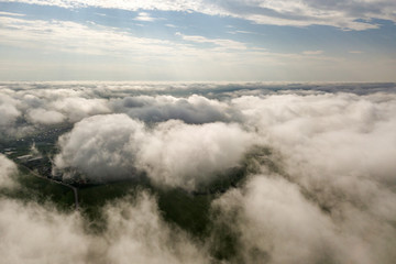 Aerial view of white clouds above a town or village with rows of buildings and curvy streets between green fields in summer. Countryside landscape from above.