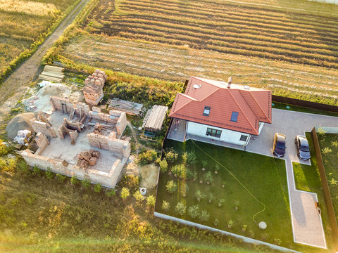 Top down aerial view of two private houses, one under construction with concrete foundament and brick walls and another finished with red tiled roof.
