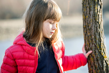 Portrait of a pretty child girl standing near a tree trunk in autumn outdoors.
