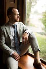 Handsome young groom sitting on window sill on wedding day