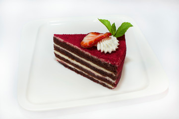 A piece of chocolate cake with white cream, garnished with red powder, strawberries, whipped cream and a sprig of mint