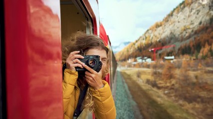 Excited happy woman, smiling and laughing leans out of train carriage window. Traveling young woman with photocamera during amazing trip on bernina express train - Powered by Adobe