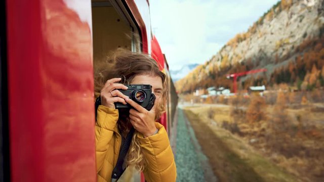 Excited happy woman, smiling and laughing leans out of train carriage window. Traveling young woman with photocamera during amazing trip on bernina express train
