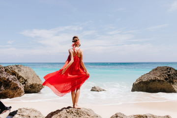 Full-length photo from back of pretty female model standing on big stone on ocean background....
