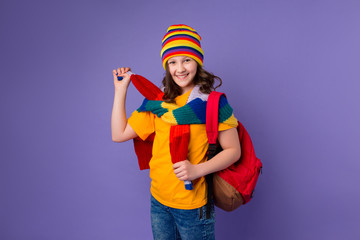 Schoolgirl girl smiles in yellow T-shirt, knitted sweater and multi-colored hat with a backpack on her shoulder on a purple background