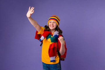 Schoolgirl girl smiles in yellow T-shirt, knitted sweater and multi-colored hat with a backpack on her shoulder on a purple background
