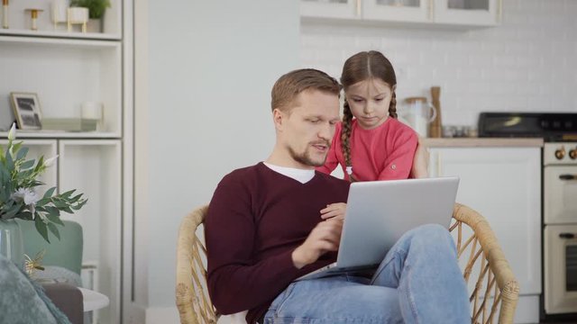 Little Girl Running To Father Sitting In Armchair At Home And Working On Laptop Computer. Kid Embracing Parent And Listening To Him Explaining His Work