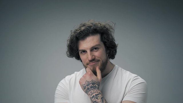 Long haired bearded young fit guy in white t-shirt posing on camera. Isolated on white touching his chin with tattooed arm and looking straight to camera.