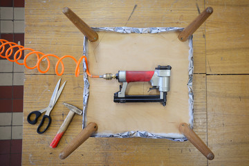 Upholstery workshop. Table top view. Work on upholstered chair seat. Pneumatic stapler with...