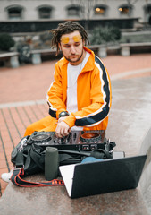 Fototapeta na wymiar Fashionable DJ man in stylish casual clothes sits on the street with a DJ controller and a laptop and plays music networks on a column. Stylish DJ works with DJ mixer outdoors