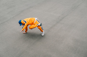 A bright dancer in an orange tracksuit shows a dance break performance on a paved playground, isolated. Professional hip hop dancer dancing on the street. Background. Copy space. Top view.