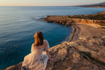 A beautiful young woman in pink dress sits at the edge of cliff and feeling sea breeze at her relaxing face. Sunset by the sea.
