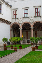 Courtyard of a historic castle complex in Trento, Italy. Trees in pots mark the way. Tourists walk among the colonnades.
