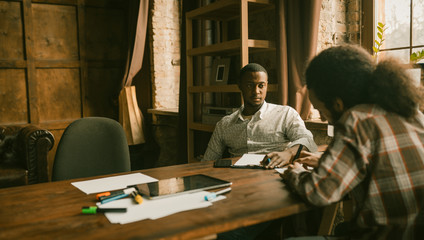 Two Young Businessmen Working In Loft Style Interior, Successful African Entrepreneur Is Thoughtfully Listening To His Arab Colleague While Sitting At Wooden Desk In Office