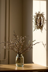 a glass jar of water with a bouquet of willow branches with fluffy buds on a wooden table in the kitchen, a wreath of willows hanging on the wall, sunlight, hard shadows