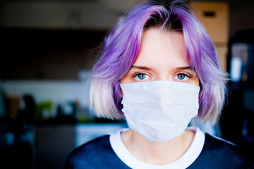 Young woman in a medical mask looks to the camera inside her house.