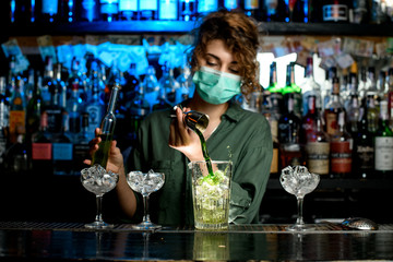 girl bartender in medical mask pours green liquid into glass with ice using beaker.