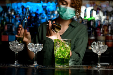Close-up girl bartender in medical mask carefully pours green liquid into glass with ice using beaker.
