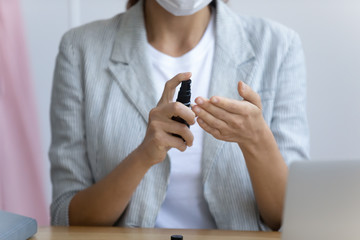 Woman sit at desk clean hands uses alcohol-based sanitizer spray to decrease infectious agents on arms protecting herself during COVID-19 pandemic infectious disease outbreak close up conceptual image