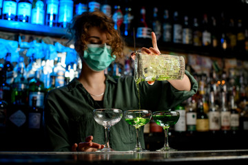 Young woman bartender in medical mask carefully pours drink from large glass into wineglasses.