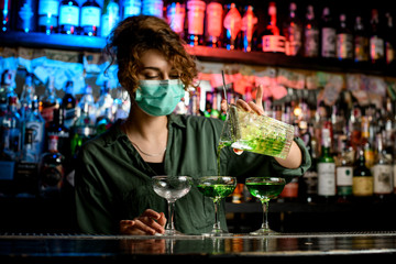 Young woman bartender in medical mask professionally pours drink from large glass into wineglasses.