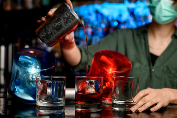 Close-up bartender girl in medical mask pour drink from steel glass into glasses with ice.