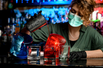 bartender girl in medical mask and black gloves accurate pour drink from steel glass into glasses with ice.