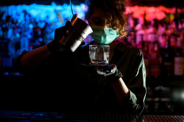 Young barman girl in medical mask and black gloves accurate pour drink from steel glass into glasse...