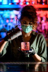 Woman barman in medical mask and black gloves holds glass and focused pours drink into it.