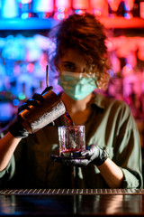 Woman barman in medical mask and black gloves holds glass and attentively pours drink into it.