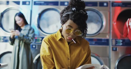 African American young beautiful girl in yellow glasses sitting in laundry service room. Woman with lollypop reading magazine while waiting for clothes to be washed. Female clients on background.