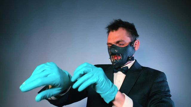 A man in a mask on his face puts gloves on his hands, straightens clothes and makes a movement of his hands into the camera. Kinky party invitation concept.