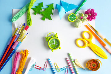 A green alarm clock on a blue table surrounded by pencils, paints, threads, colored paper and other stationery. Children's creativity. The concept of creativity for children on vacation, homework