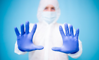 A male doctor in a white virus-protected suit and blue medical rubber gloves with a mask on his face holds out in front of his hand showing them. The doctor on the background blurred background.