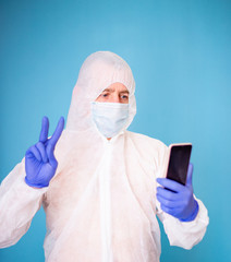 A virologist in a white mask and blue rubber gloves is dressed in a viral protective suit talking on the phone in a video call.
