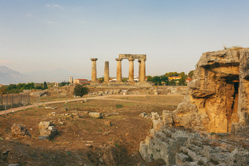 Ruins of Temple of Apollo in Corinth Greece standing up on a hill with remenants of rock walls scattered about under a bright sun with mountians and a blue sky behind