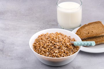 Cooked buckwheat in white bowl with spoon, glass of milk, piece of bread on grey background. Close up, space for text, horizontal format.