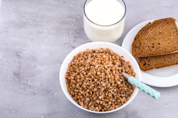 Cooked buckwheat in white bowl with spoon, glass of milk, piece of bread on grey background. Close up, space for text, horizontal format.