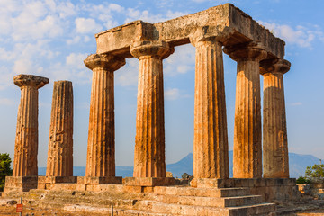 Obraz premium Ruins of Temple of Apollo in Corinth Greece standing up on a hill with remenants of rock walls scattered about under a bright sun with mountians and a blue sky behind