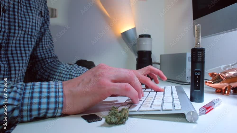 Poster designer sitting at a desk typing text on a computer keyboard. smokes a vaporizer with cannabis. the