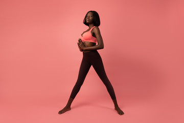 Fototapeta na wymiar Full lenght photo of a sporty african girl in sportswear stands with wide legs, holding hands together and looking seriuosly at the camera, isolated over pink background