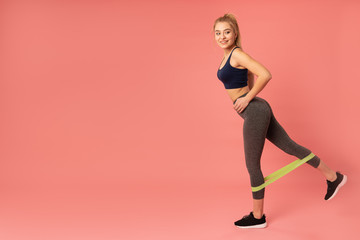 Indoor picture of athletic adorable model keeping her hands on her waist, looking directly at camera, sporty girl having elastic band for fitness, training legs, wearing grey combidress
