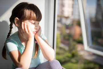 Adorable sad child girl by the window at home. Quarantine. Coronavirus. Epidemic.