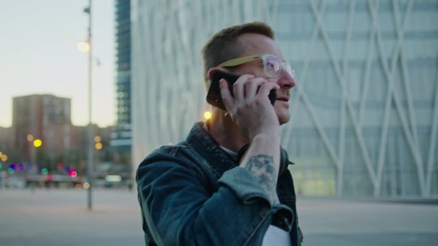 Handsome Man Walking and Answering Call. Good-Looking Male Person Wearing Glasses and Headphones Talking on Phone in Street. Online Communication Concept. Slow Motion Arc Tracking Shot