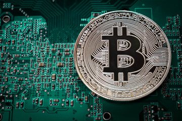 A silver bitcoin coin lies on a green printed circuit board.